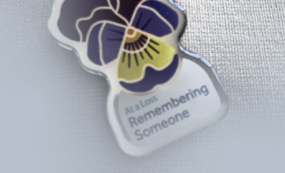 A close-up of a pin featuring a stylized flower design in blue, purple, and yellow. The pin has the words At a Loss Remembering Someone written on it against a white background. The image is set on a textured surface.