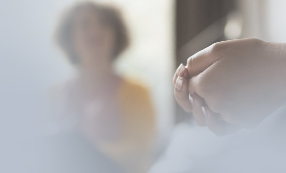 Close-up of a persons hands clasped together in the foreground, with a blurred background showing a second person sitting. The scene has a soft, muted color palette, creating a gentle and serene atmosphere.