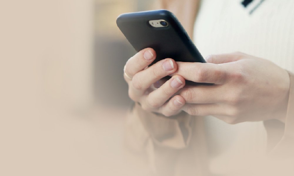 Close-up of a persons hands holding a smartphone. The person is wearing a light sweater and a brown coat. The background is softly blurred, emphasizing the focus on the hands and phone.