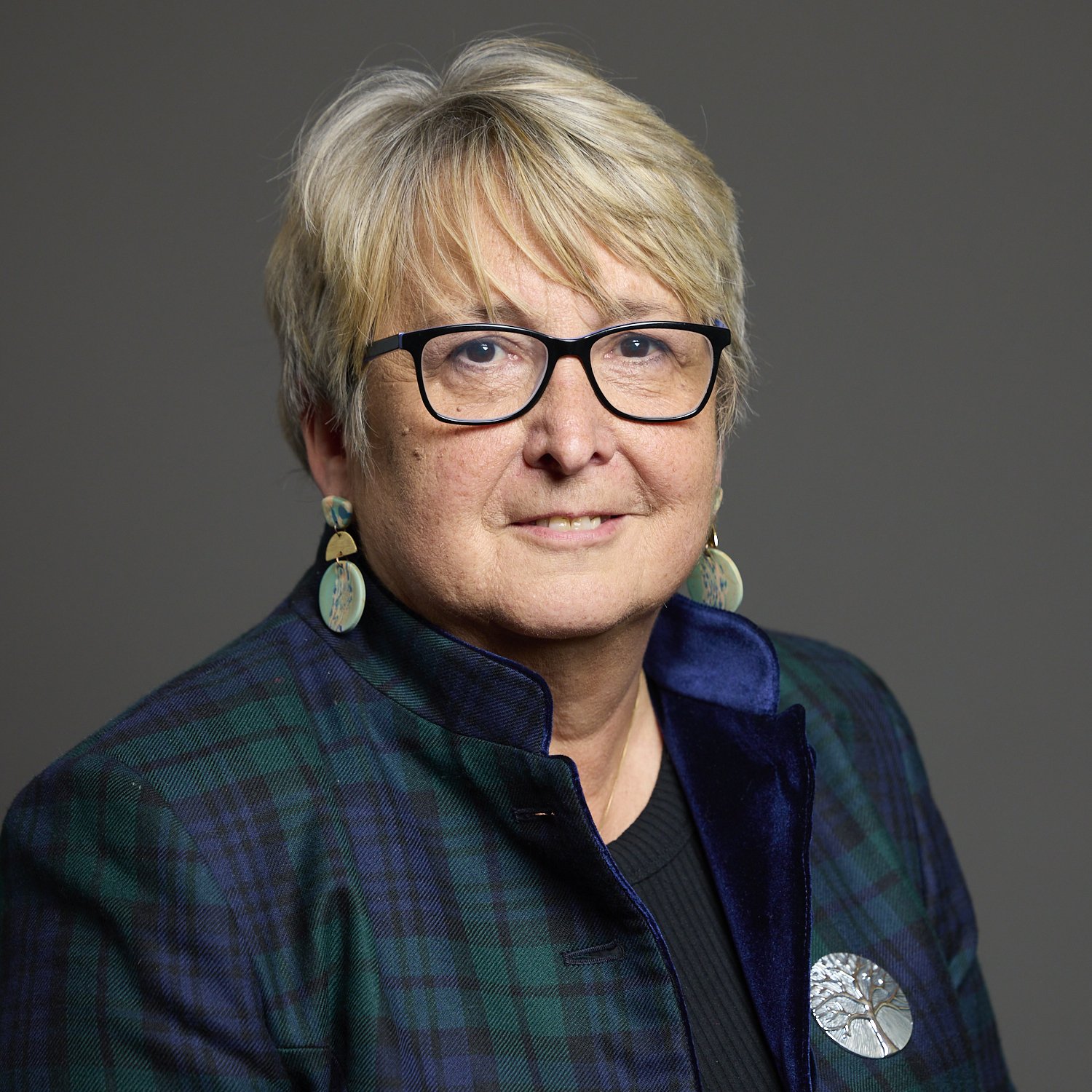 An older woman with short gray hair and glasses wearing a plaid jacket, a dark shirt, and round earrings, standing against a neutral background.