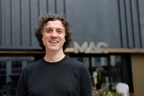 Smiling person with curly hair wearing a black shirt, standing in front of a building with 'MAC' sign visible.