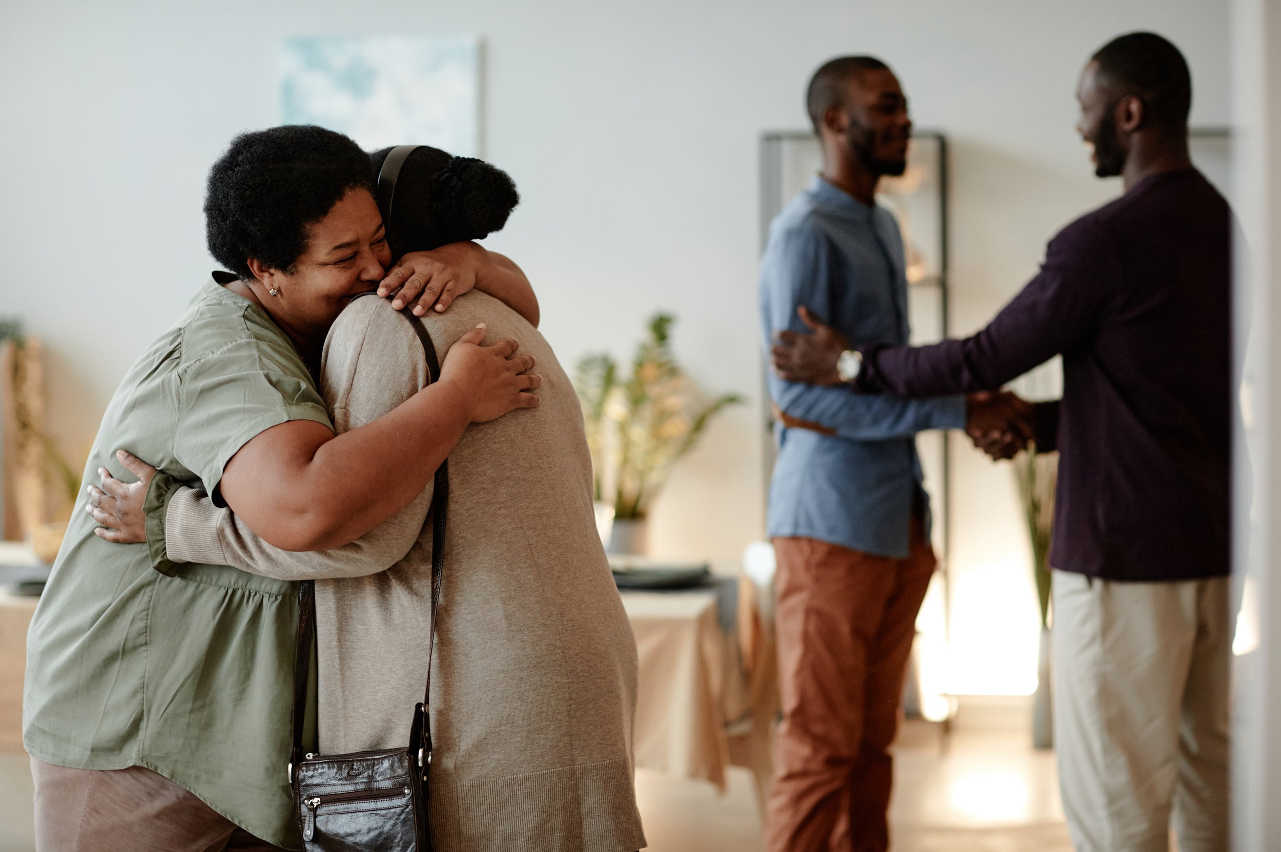 Two people hugging joyfully in the foreground, and two others greeting each other with a handshake in the background, indoors with a casual setting.