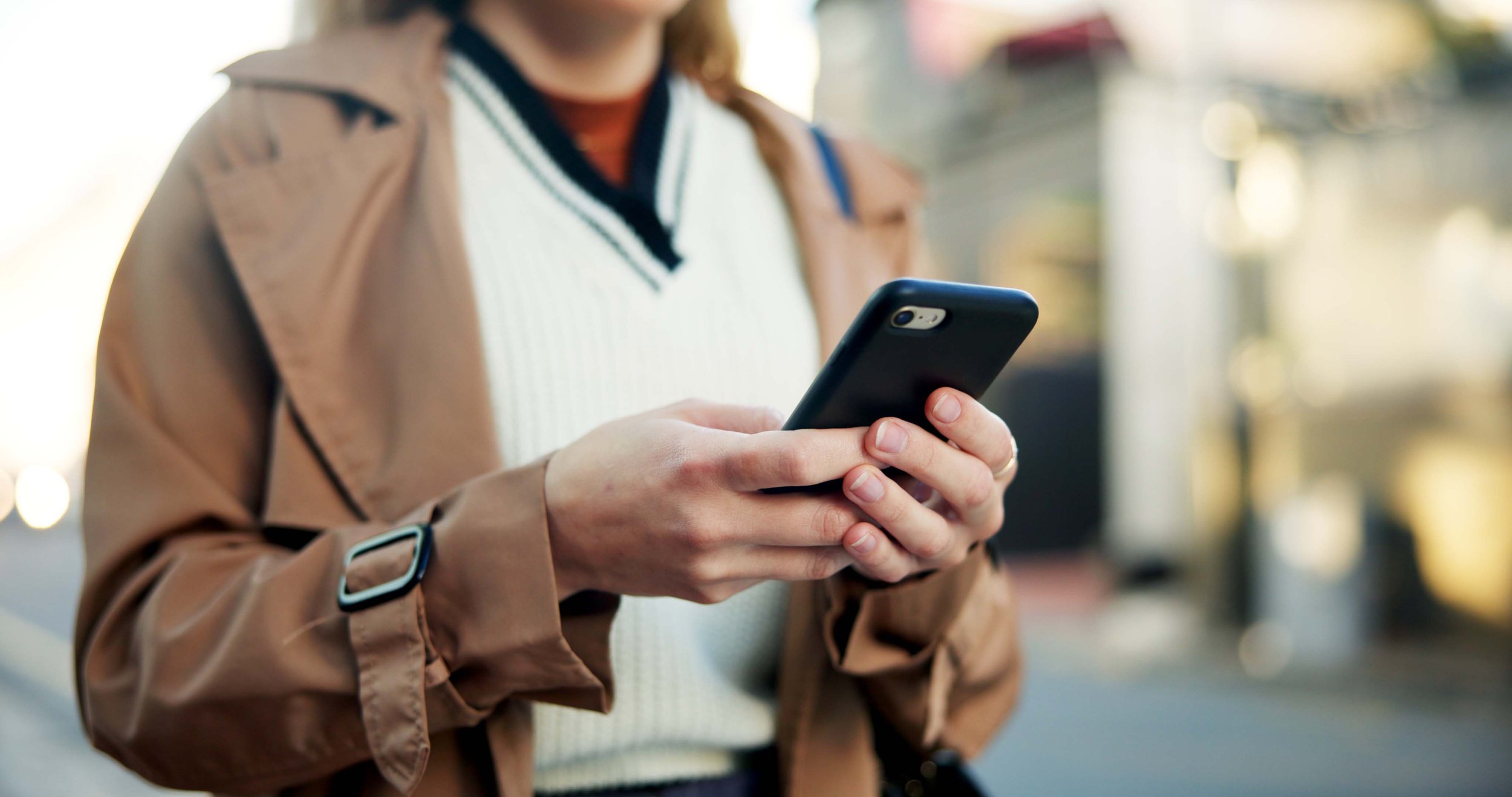 Person wearing a brown coat using a smartphone outdoors, focus on hands.