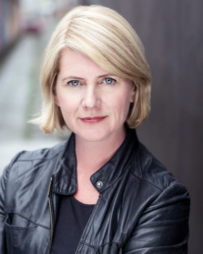 Portrait of a woman with short blonde hair in a black leather jacket, standing in an outdoor setting.