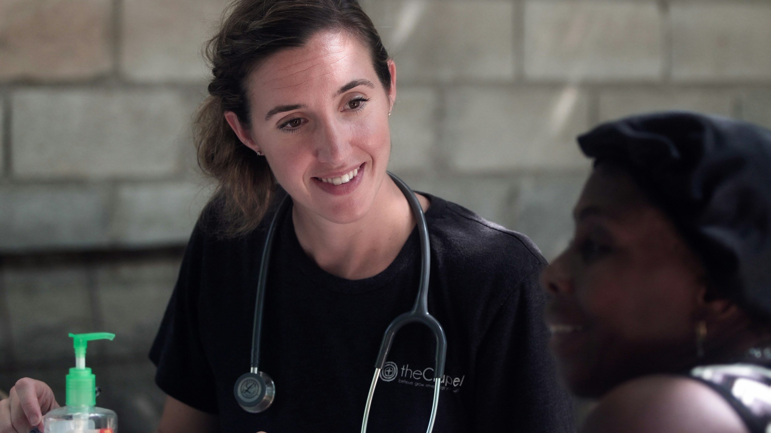 A female doctor in a black shirt engages in conversation with a person in a public health setting.